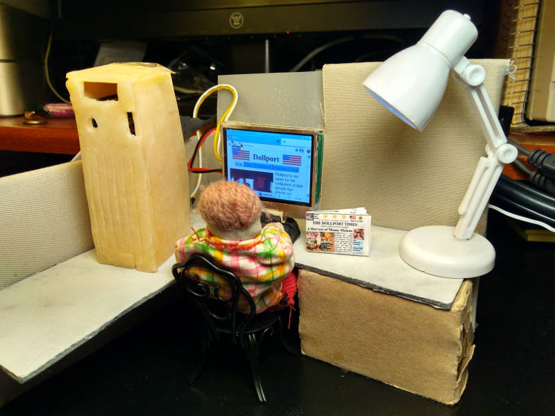 A doll computer sits in a doll cubicle both serving and browsing a doll website